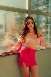 Fashionable woman in pink ensemble with heart-shaped sunglasses by a bright window, exuding confidence.