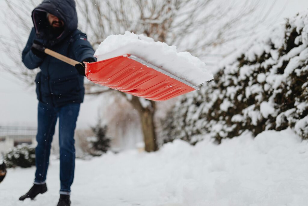 A man uses a red shovel to clear snow in a snowy yard, emphasizing winter maintenance.