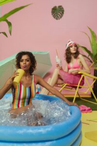 Two women enjoying a vibrant indoor poolside setup with drinks in hand, exuding summertime joy.