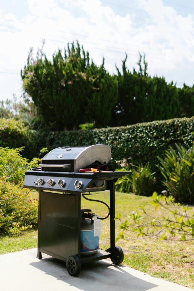 A sleek gas grill on a sunny day surrounded by green foliage and trees.
